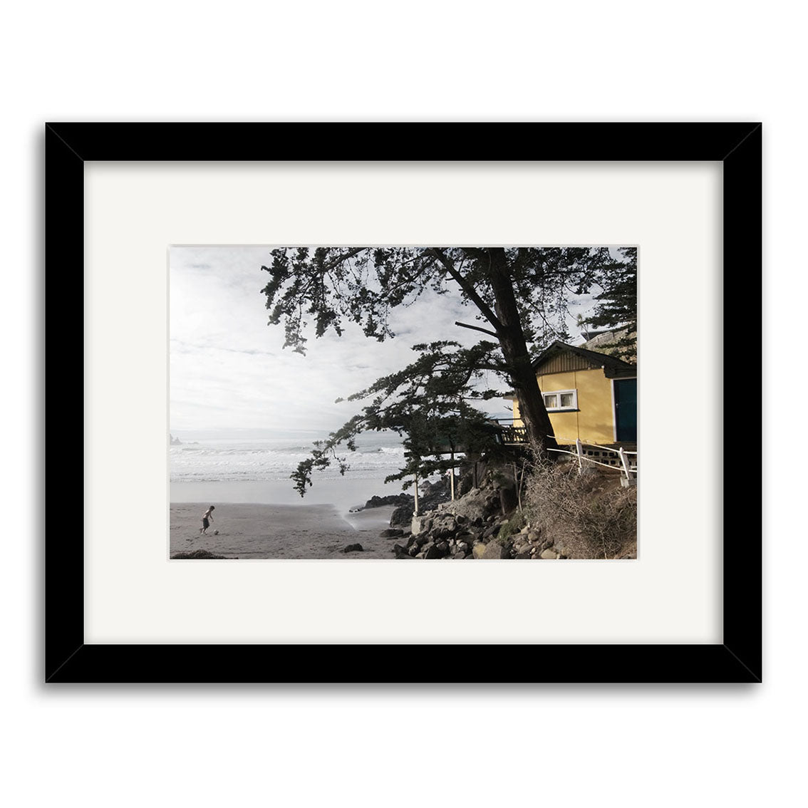 A person walks on a sandy beach near the ocean, with a yellow house partially hidden by trees. This scene is beautifully captured in the Taylors Mistake - RP175 Reuben Price print, elegantly framed with a black and white mat and frame.