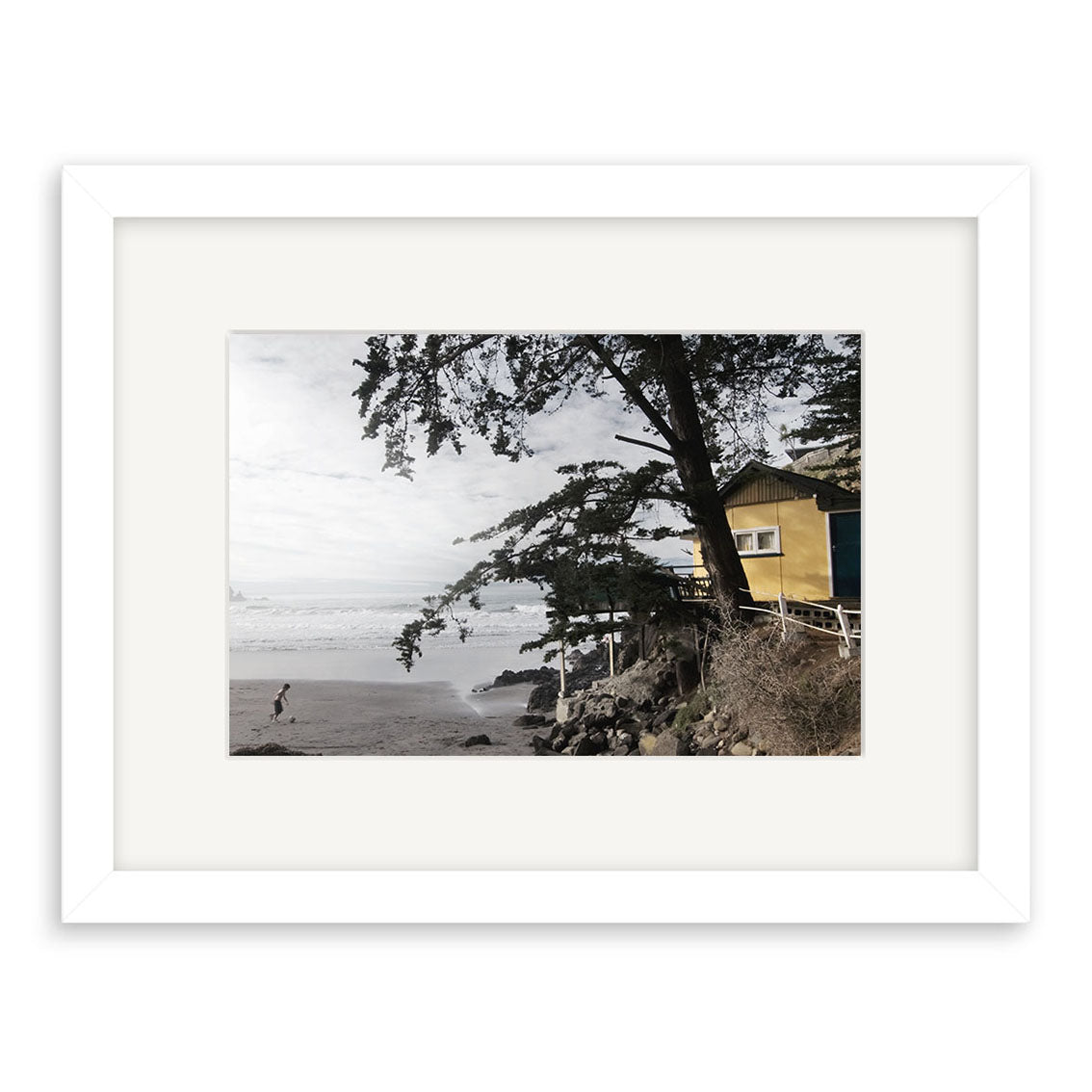A framed photo titled Taylors Mistake - RP175 features a sandy beach, one person walking by the shoreline, a large angled tree, and a yellow house on a rocky bluff—an exquisite New Zealand photographic print.