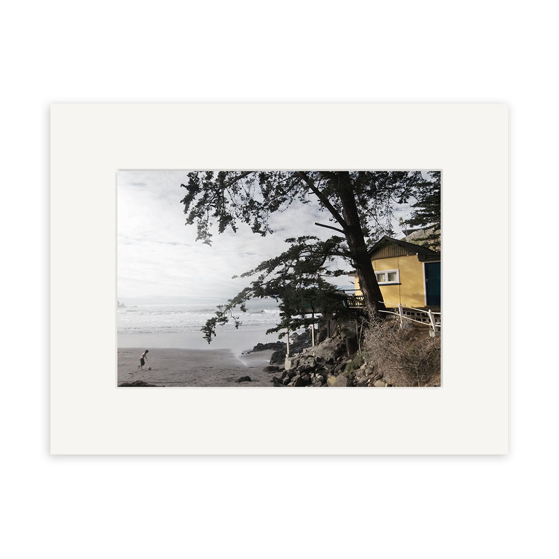 A person walks on a sandy beach near the calm ocean, with a large tree leaning over a yellow house on the rocky shoreline. Cloudy skies evoke the tranquil beauty of New Zealand in Taylors Mistake - RP175 photographic print.