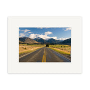 Arthurs Pass - RP177 depicts a straight, two-lane road with a yellow centerline leading to mountains in New Zealand, bordered by grassy fields. The photo is mounted with a white mat on archival quality matte paper.