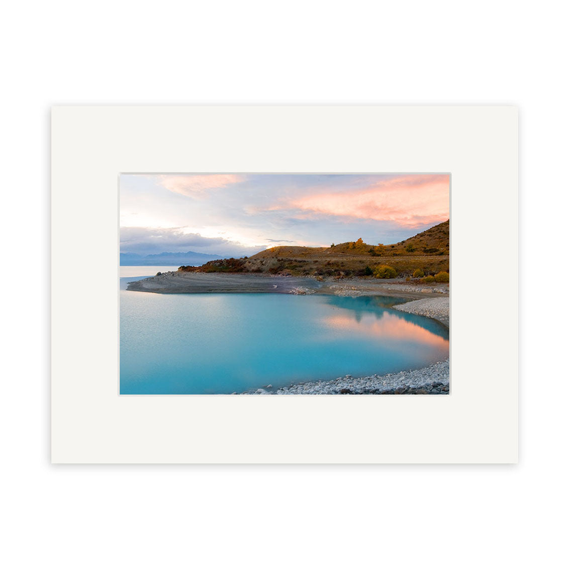 “Lake Pukaki - RP178” by Reuben Price features a tranquil turquoise lake, rocky shore, and sunset-lit hills under pink-orange clouds with a white mat border, part of the New Zealand photographic prints on archival matte paper.