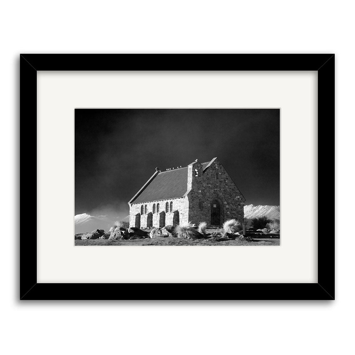 Lake Tekapo - RP179: Black and white photo of a small stone church with a dark roof, surrounded by rocks, grass, and distant mountains. Presented in a black frame with white mat on archival quality matte paper, capturing New Zealand’s beauty.