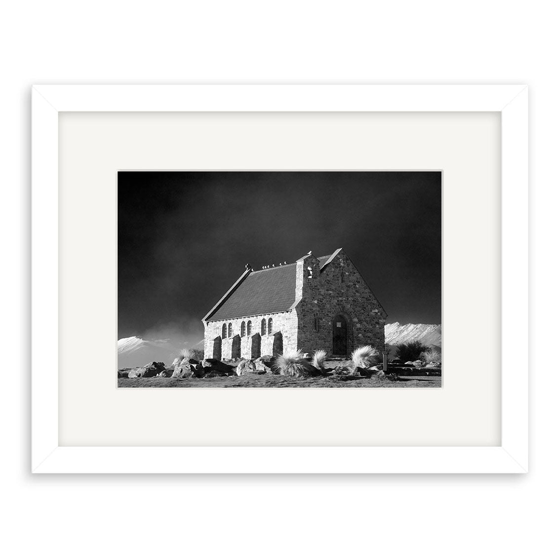 Lake Tekapo - RP179: Black and white photo of a small stone church with steep roof in New Zealand’s rocky landscape, dramatic dark sky, printed on archival matte paper and framed with a white mat and frame.