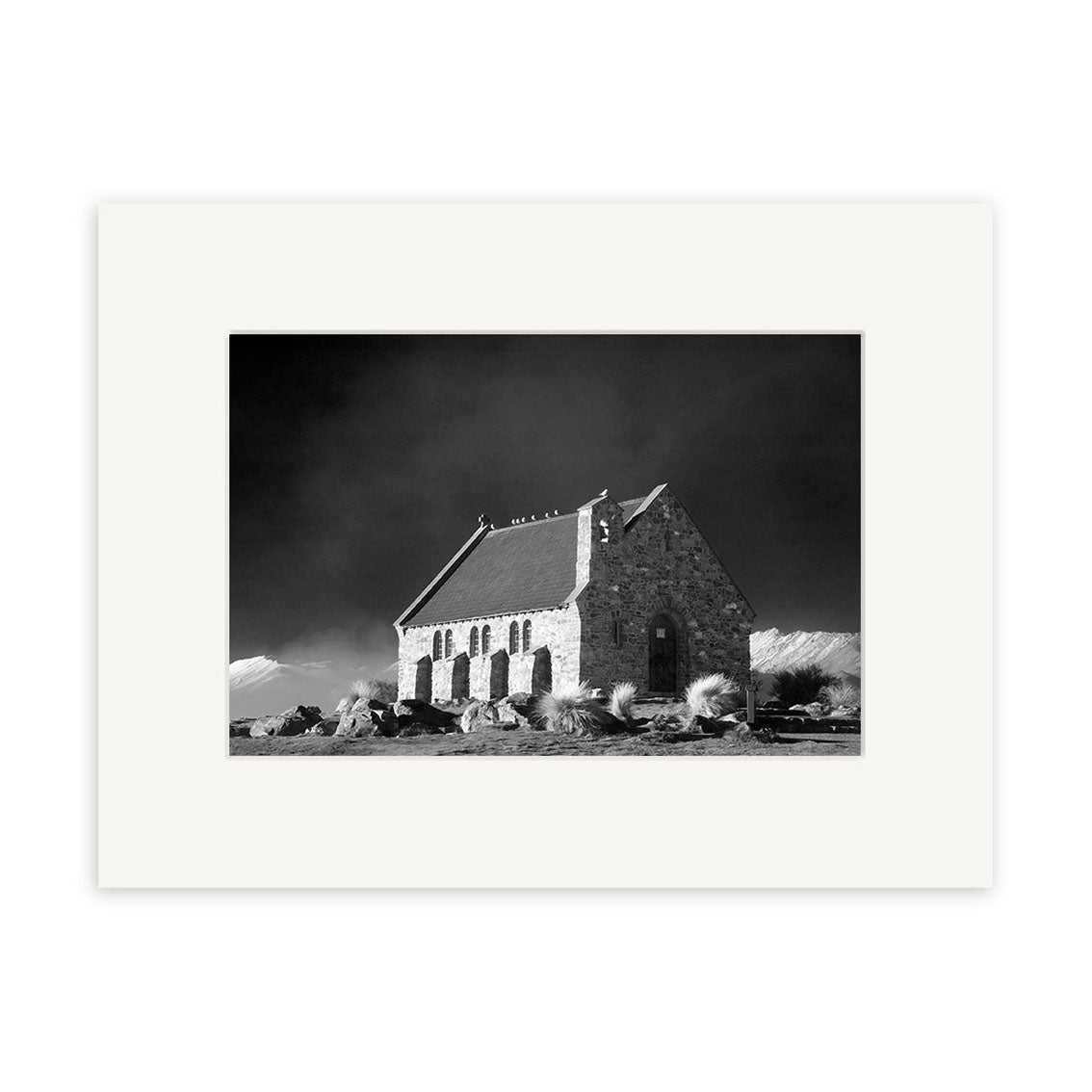 Lake Tekapo - RP179: Black and white photo of a small stone church in New Zealand with arched windows and steep roof, set against a dramatic sky. Surrounded by rocks and grass, image is matted with archival quality matte paper.