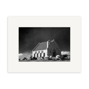 Lake Tekapo - RP179: Black and white photo of a small stone church in New Zealand with arched windows and steep roof, set against a dramatic sky. Surrounded by rocks and grass, image is matted with archival quality matte paper.