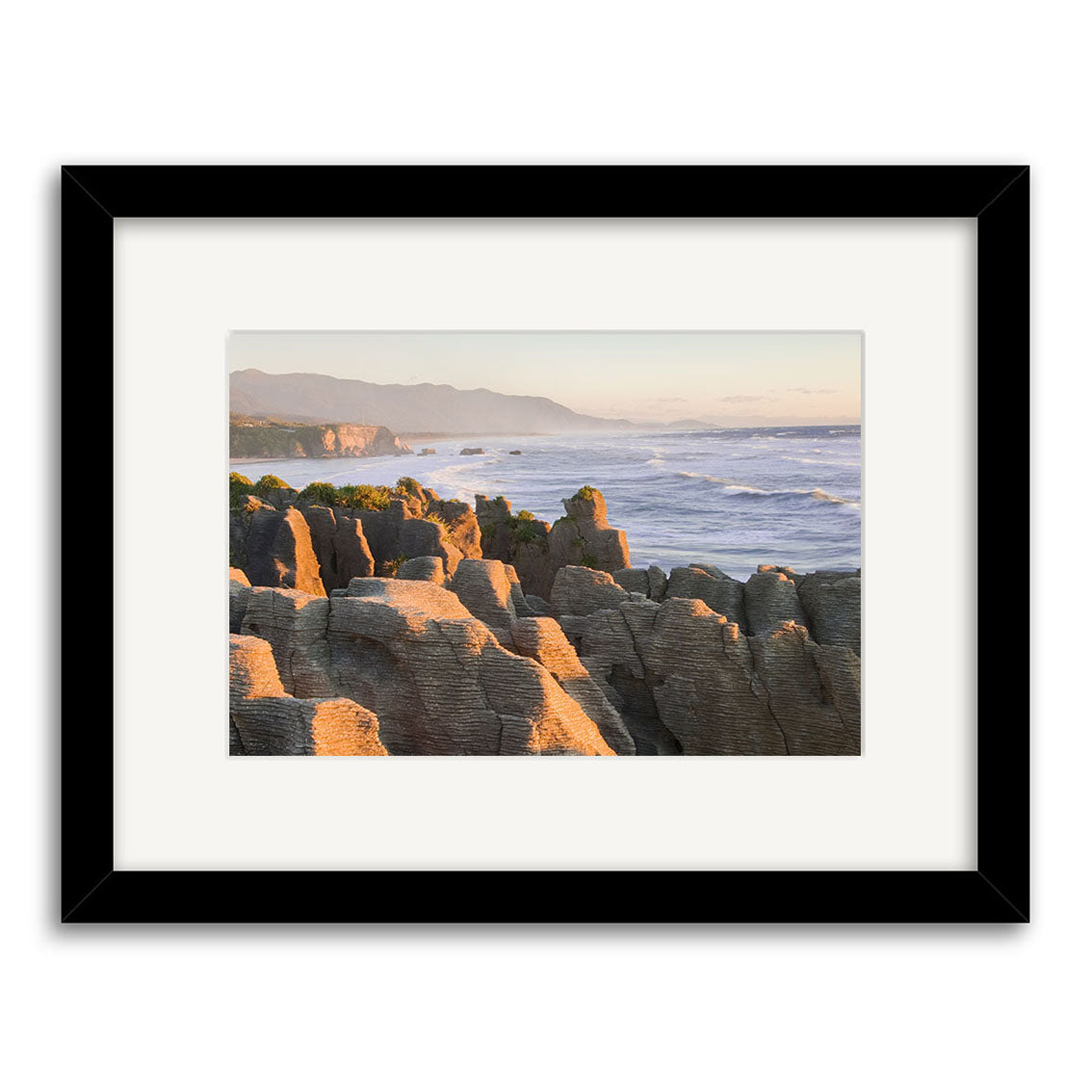 Punakaiki Sunset - RP180: Framed photograph of coastal rock formations under soft sunlight, waves crashing with distant mountains and a pale sky. Printed on archival-quality matte paper, ideal for photographic prints.