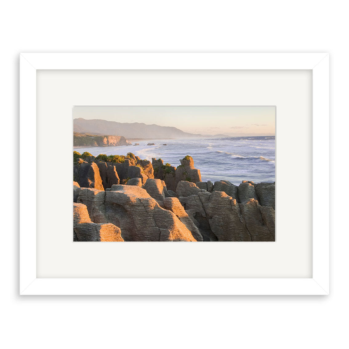 Punakaiki Sunset - RP180: A framed archival matte print featuring layered rock formations by the New Zealand ocean, waves, and distant mountains at sunset. Displayed with a white mat and frame.