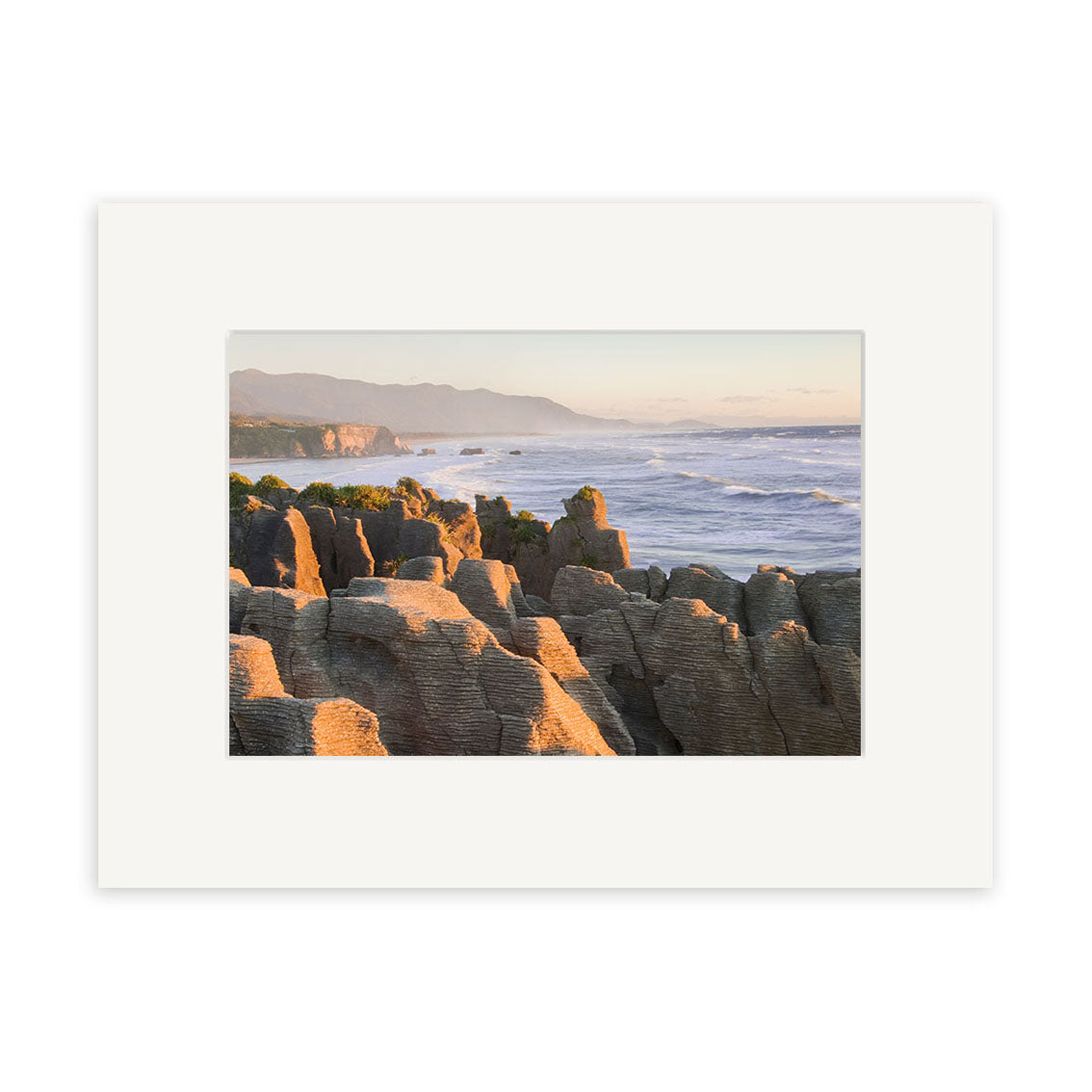 Punakaiki Sunset - RP180 captures New Zealand’s coastal landscape with layered rocks, ocean waves, and distant mountains under a clear sky in golden light. Available as archival quality matte paper photographic prints.