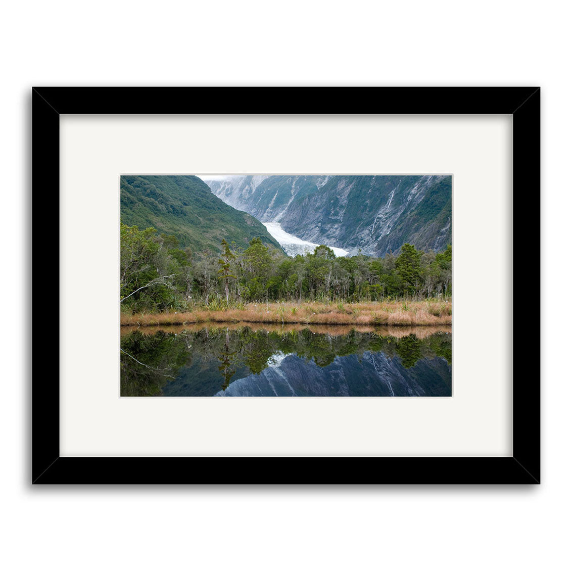 Franz Josef Glacier - RP181: A framed photo print of a glacier nestled between mountains in New Zealand, with lush forest and brown grass reflected in tranquil water, printed on archival-quality matte paper.
