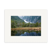 Franz Josef Glacier - RP181: A landscape photo of a glacier between green mountains in New Zealand, with trees and grass reflected in calm water. Printed on archival matte paper, bordered by a white matte frame.