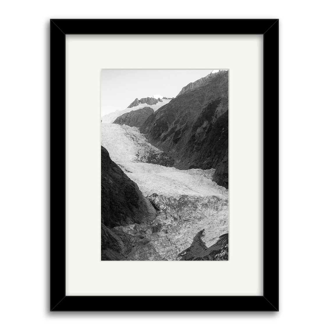 The Franz Josef Glacier 1 - RP183 is a black and white photo print of a glacier flowing between New Zealand’s rocky slopes, shown in a black frame with white mat and printed on archival quality matte paper, highlighting textured ice crevasses.