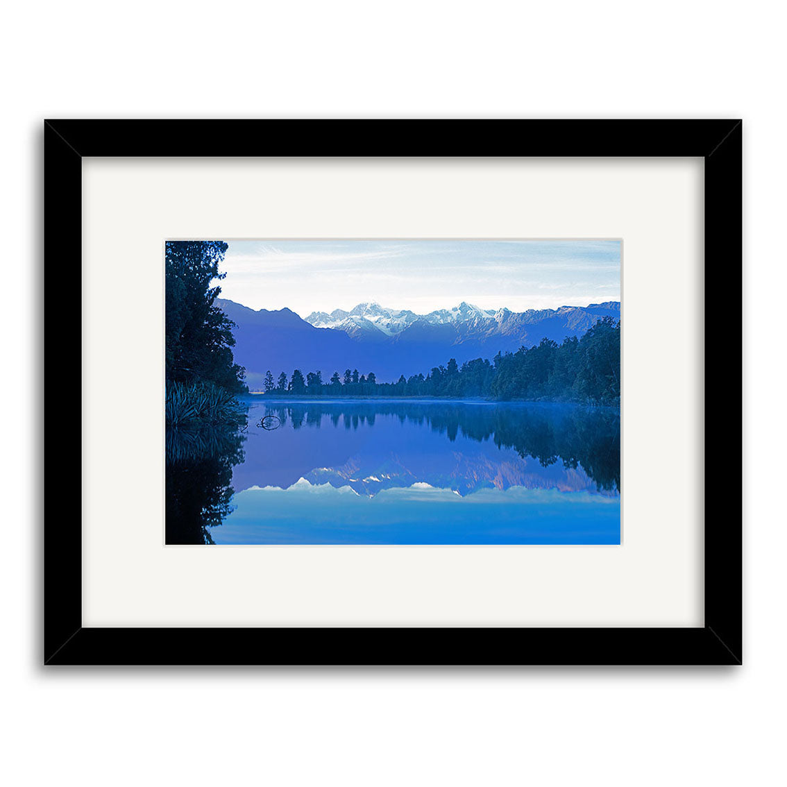 Lake Matheson 1 - RP184: Framed Reuben Price photographic print of a tranquil New Zealand lake mirroring trees and snow-capped mountains beneath a blue sky, finished with white mat and black frame.
