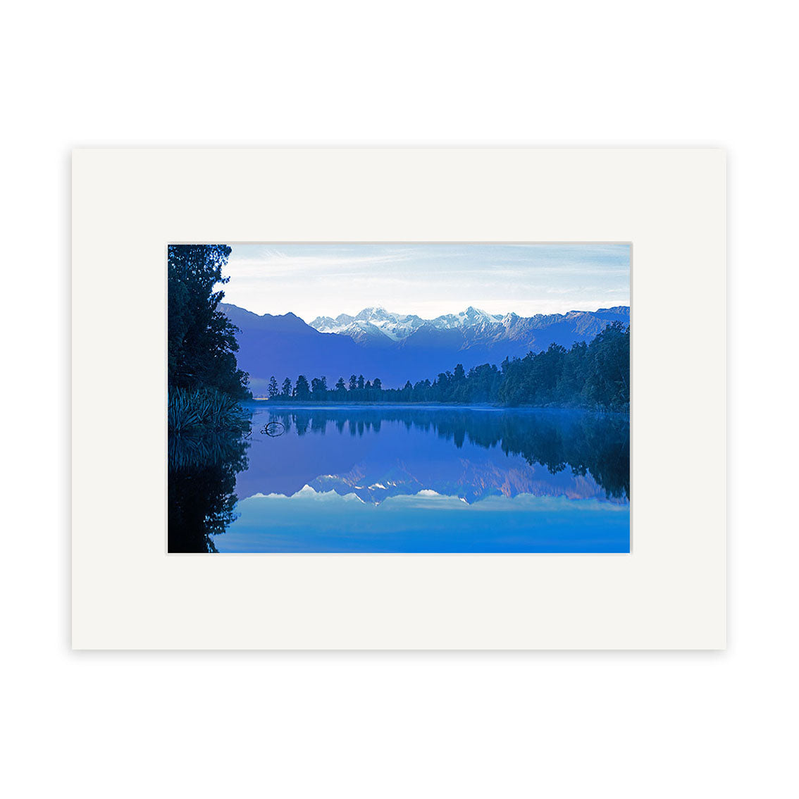 Lake Matheson 1 - RP184 by Reuben Price features a tranquil New Zealand lake reflecting trees and snow-capped mountains beneath a clear sky, bordered by a white matte frame—a stunning fine photographic print.