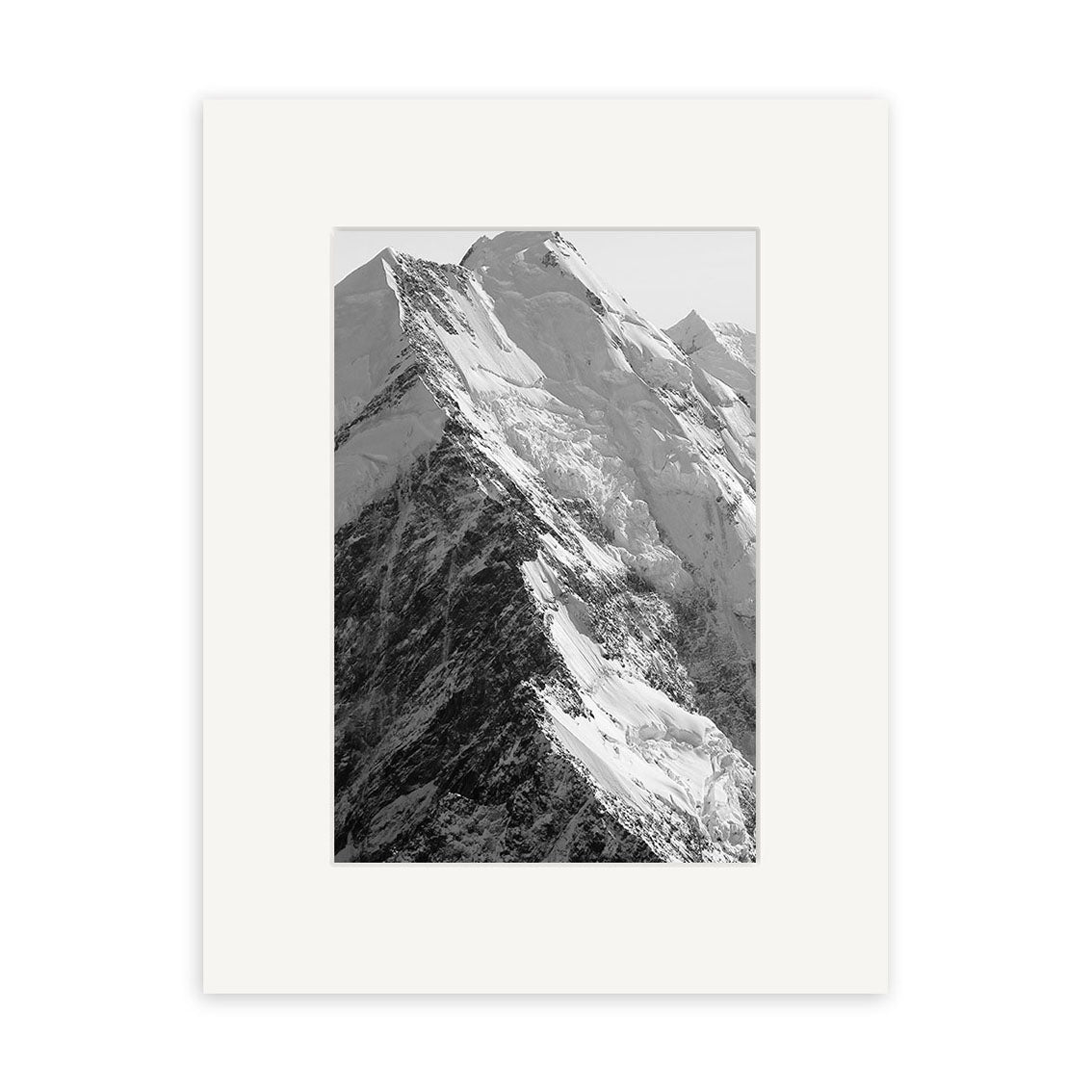 Black and white photograph Mt Cook - RP185 by Reuben Price features a snow-covered mountain peak with sharp ridges, centered in a white mat frame. This New Zealand print is created on archival quality matte paper against a plain white background.