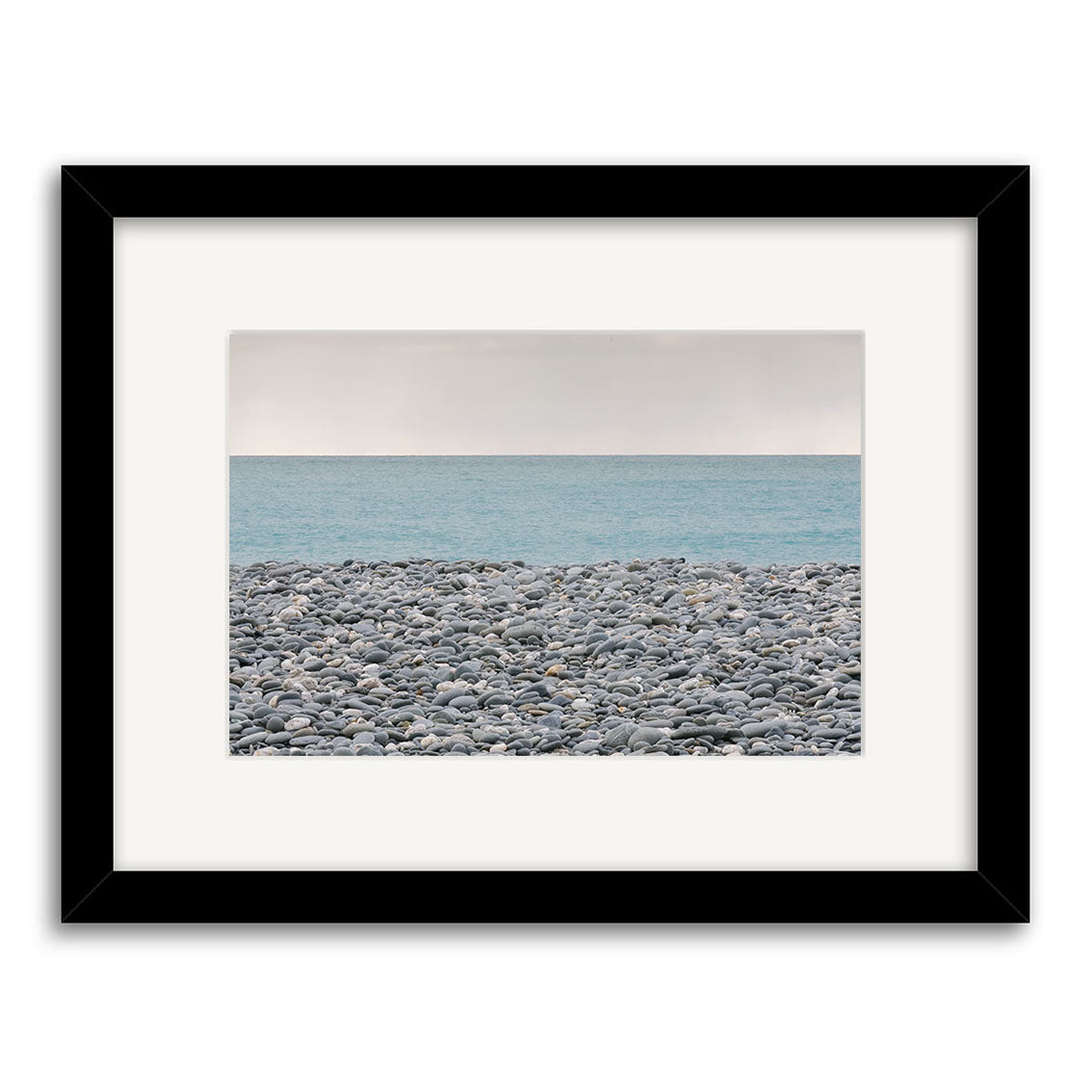 Gillespies Beach Stones - RP187: A framed photo featuring smooth grey stones on a rocky New Zealand beach, with calm blue ocean and pale sky. Centered image with white mat and black frame, printed on archival quality photographic paper.
