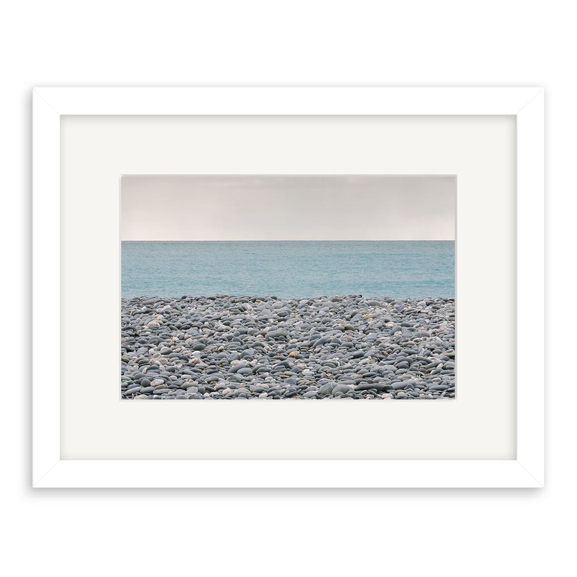 A framed, archival-quality print titled Gillespies Beach Stones - RP187 showcases smooth gray stones on a rocky New Zealand beach, with a calm blue ocean and pale sky. Classic white mat and frame complete the look.