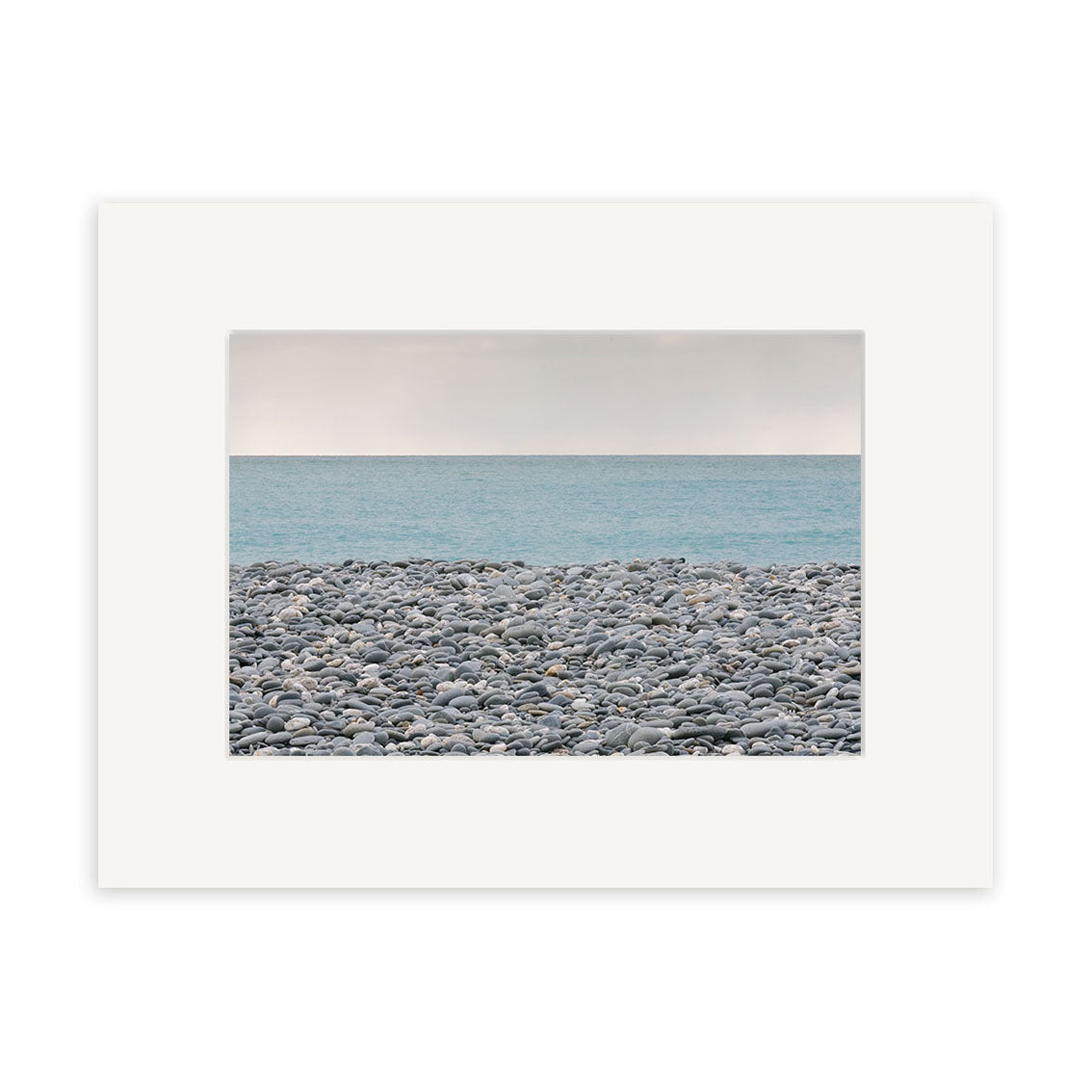 Gillespies Beach Stones - RP187 is an archival quality color photo print of a rocky New Zealand shore with smooth gray stones, calm blue sea, and pale sky, framed with a wide white mat.