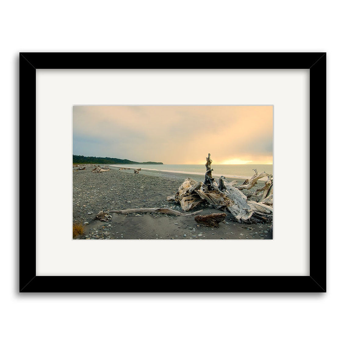 Gillespies Beach Driftwood - RP188: A photo of a rocky New Zealand beach with driftwood, calm water, and a cloudy orange-lit sky. Framed in black with white mat; printed on archival quality paper.