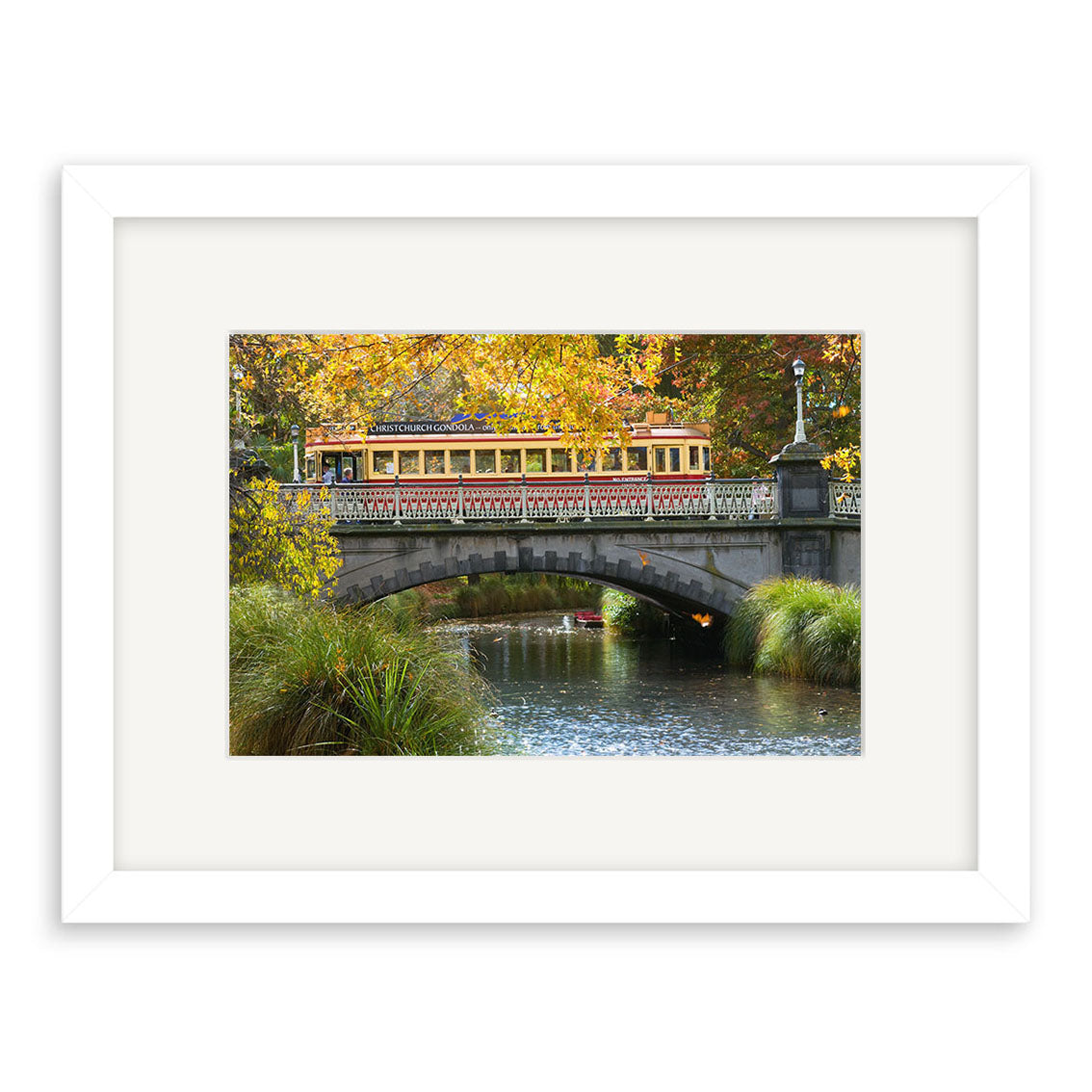 The Avon River - RP192 features a yellow and red tram crossing a stone bridge over a river in New Zealand, surrounded by autumn trees and green grass. Beautifully framed with a white mat on archival matte paper for lasting quality.