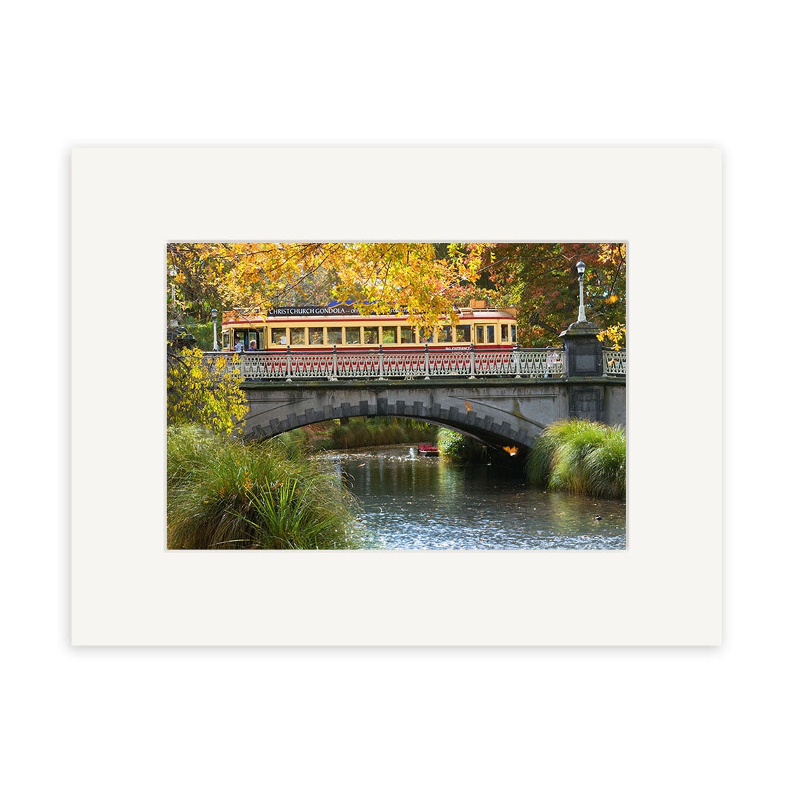 A yellow vintage tram crosses a stone bridge over the Avon River—framed by lush foliage—inspired by New Zealand, captured in vibrant photographic prints on archival quality matte paper. Product: Avon River - RP192.