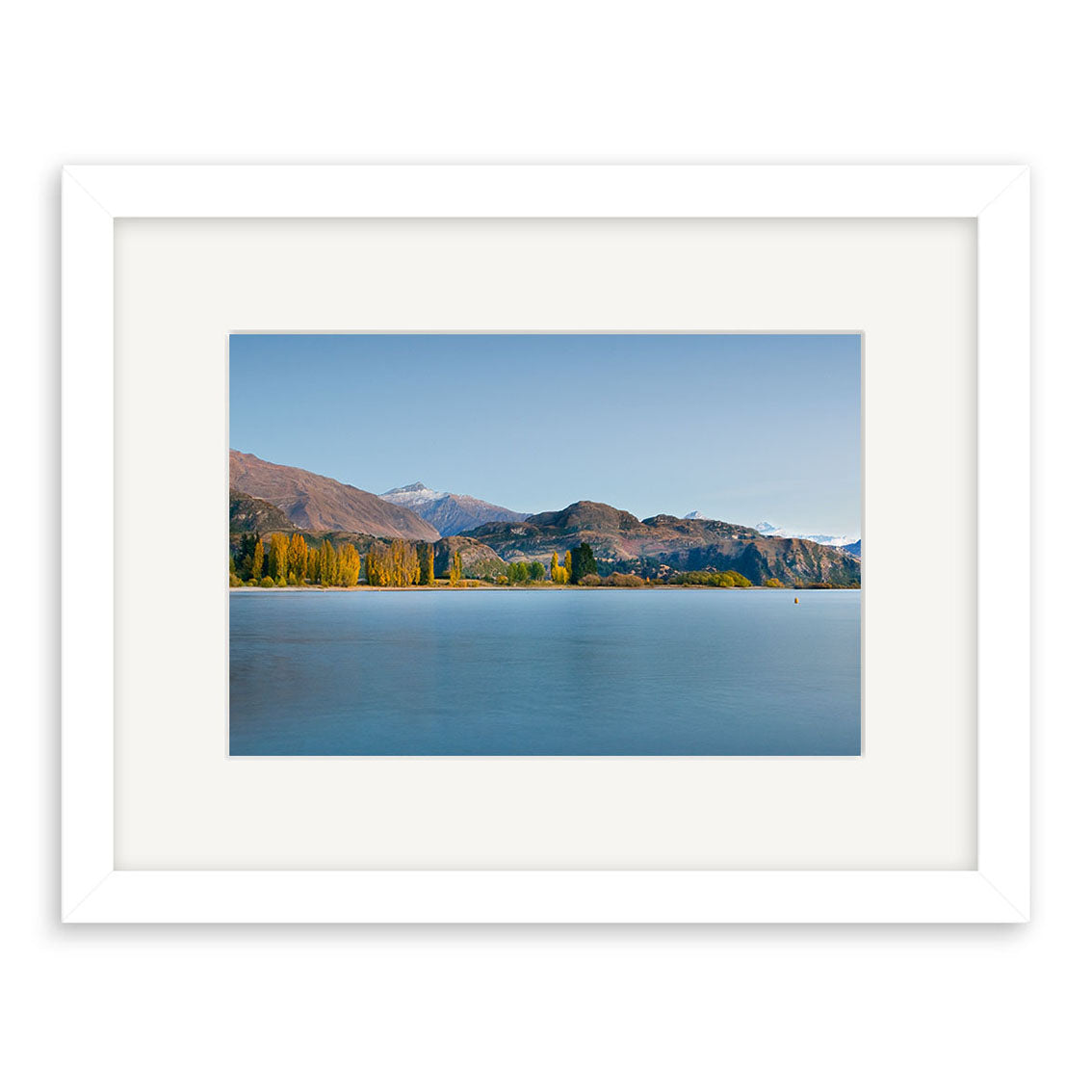 The Lake Wanaka in Autumn - RP193 is a framed photographic print showing a calm blue lake with tree-lined shores, mountains, and clear sky. Printed on archival matte paper with a white frame and wide mat border, it captures New Zealand’s serene beauty.