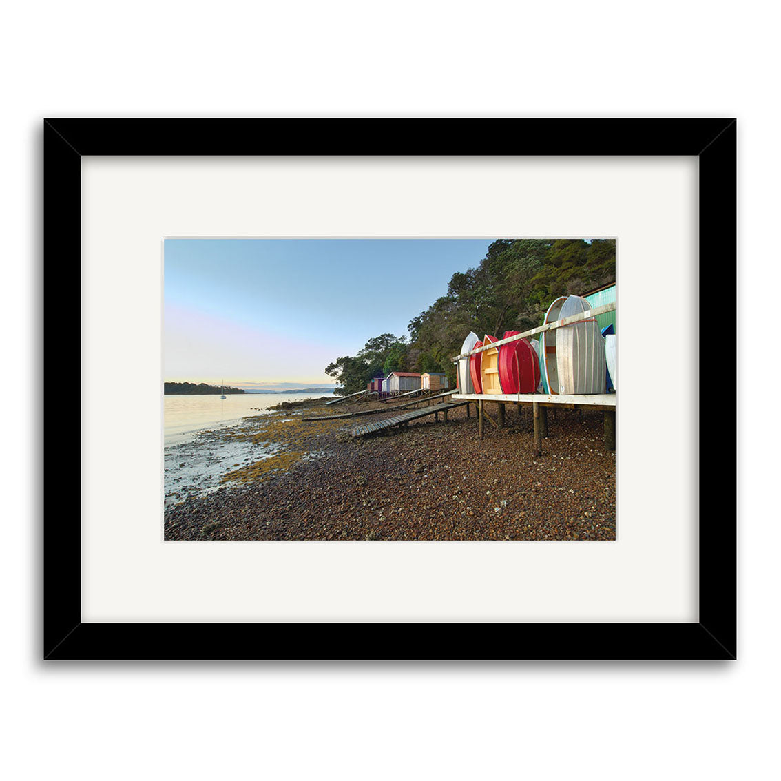 Photograph titled Rocky Bay Boatsheds - RP194 features vibrant beach huts on a pebbled New Zealand shore by calm water, under clear skies and trees. Printed on archival matte paper with a white mat in a black frame.
