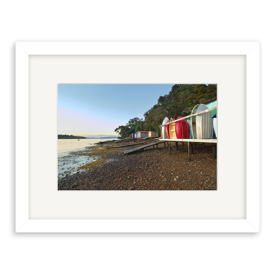 Rocky Bay Boatsheds - RP194: A white-framed print featuring small, colorful beach huts on stilts along a rocky New Zealand shore, with calm water and trees under a clear sky. Photographic print on archival quality matte paper.