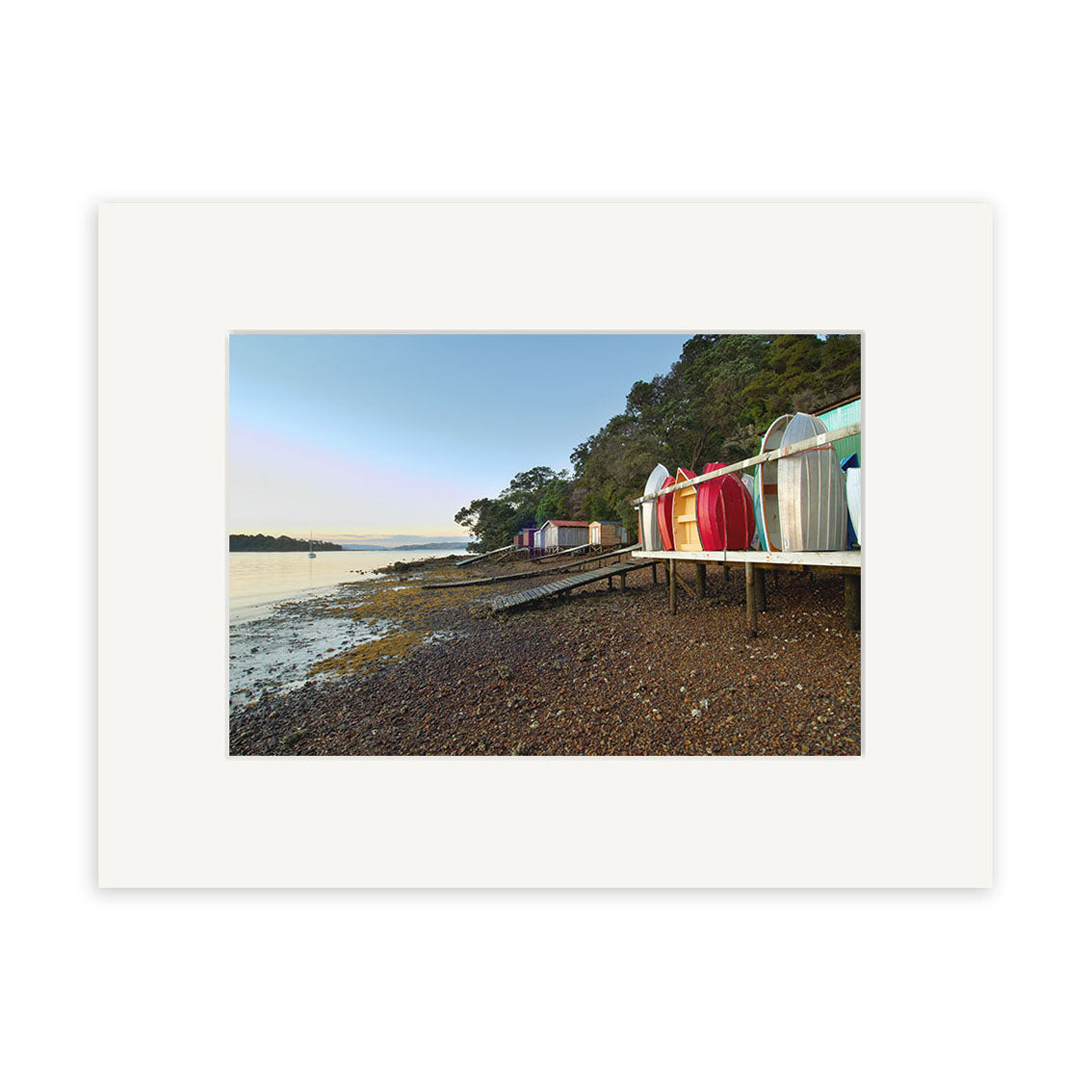Rocky Bay Boatsheds - RP194: Colorful boat sheds on a wooden platform by a rocky New Zealand shore with calm water, trees, and clear sky. Available as archival quality matte photographic prints.