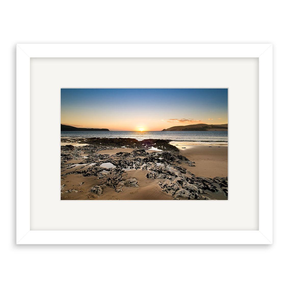 Opononi - RP197: A framed photographic print featuring a New Zealand beach landscape at sunset, with calm sea, rocky shoreline, sandy beach, distant hills, and the sun setting under a clear sky.