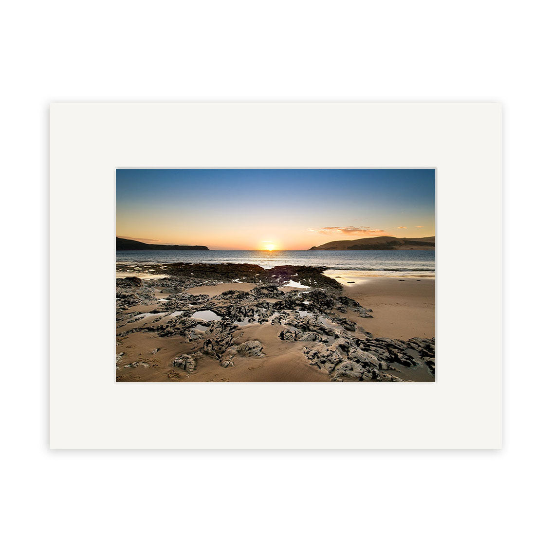 The Opononi - RP197 is a color photo of a sandy New Zealand beach at sunset with rocks, calm water, and distant hills. Surrounded by a white mat border, this archival quality photographic print ensures lasting beauty.
