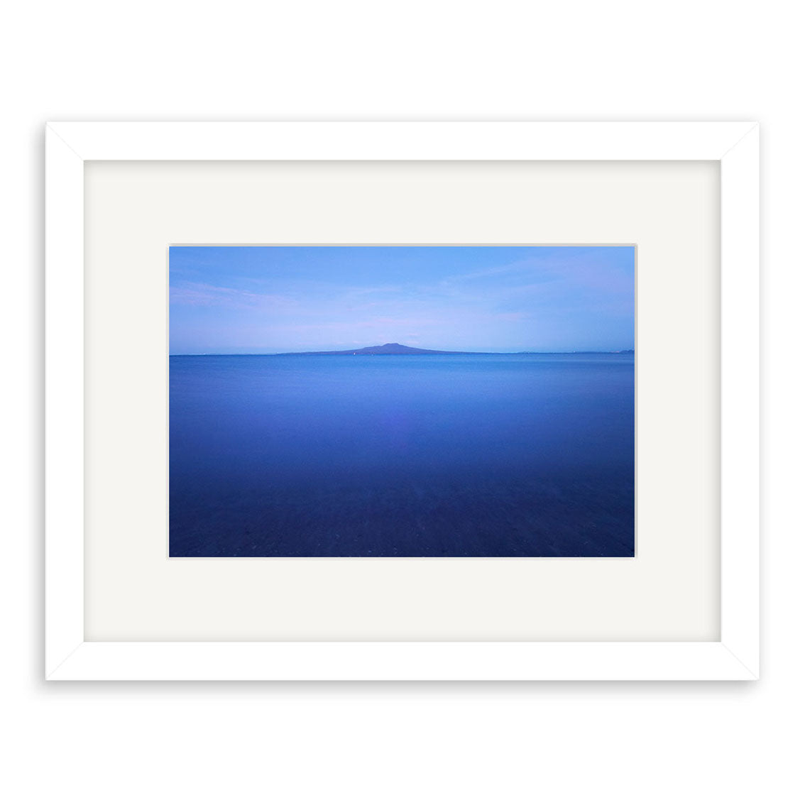 Rangitoto in moonlight - RP198: A framed photographic print featuring a tranquil blue seascape with a distant island under a pale sky. The white frame and wide mat accentuate this archival image, inspired by New Zealand’s peaceful coastlines.