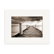 Magazine Bay - RP199: A sepia photo of a wooden pier with white posts stretching into calm water beneath cloudy skies, presented with a white matte frame and printed on archival quality matte paper.