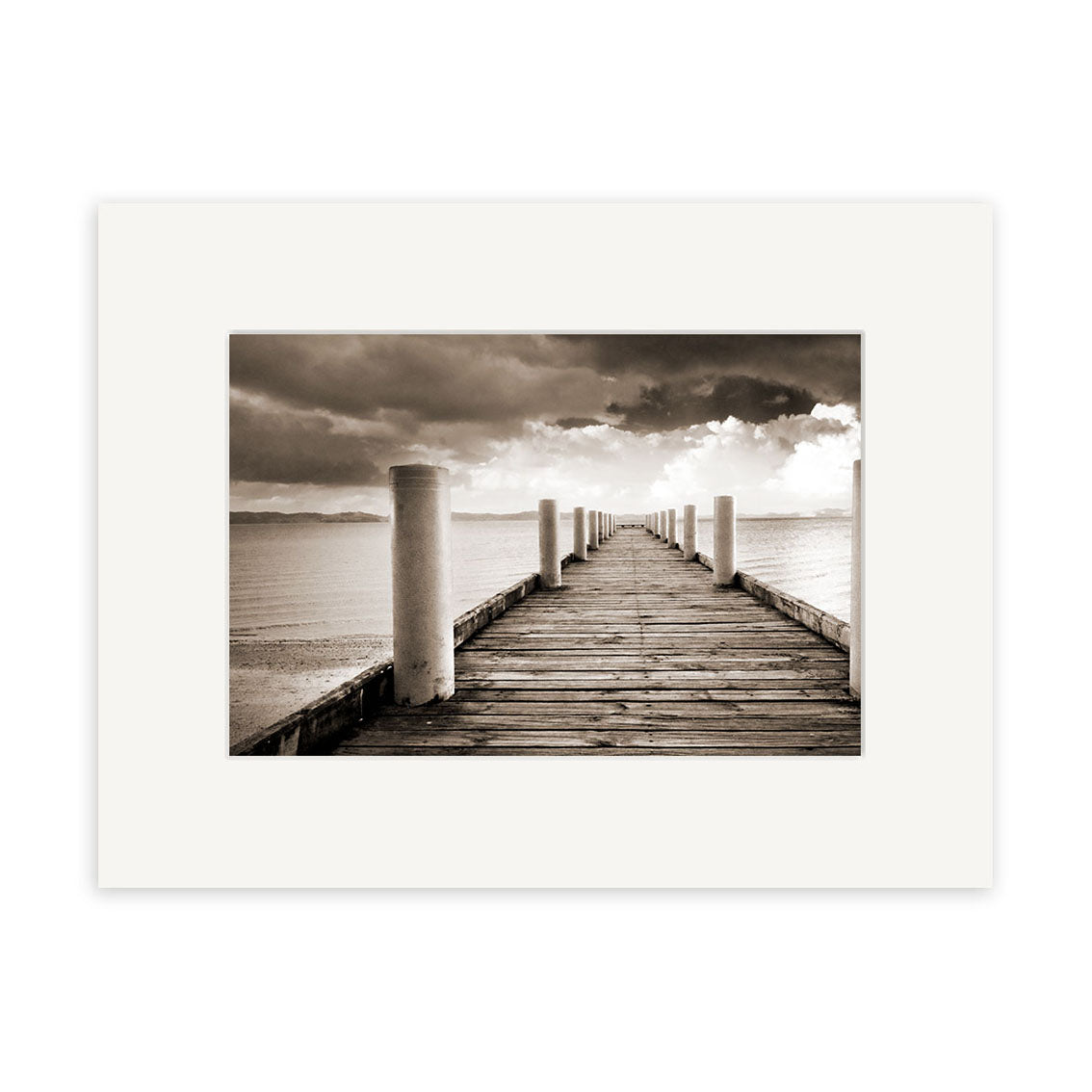 Magazine Bay - RP199: A sepia photo of a wooden pier with white posts stretching into calm water beneath cloudy skies, presented with a white matte frame and printed on archival quality matte paper.