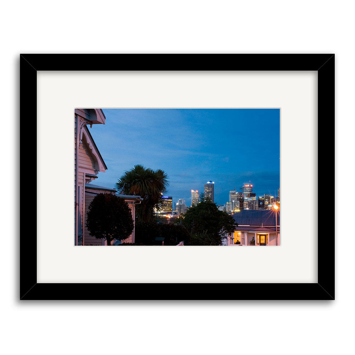 The Ponsonby - RP200 is a framed photographic print of a suburban dusk scene with houses, trees, and a city skyline on archival matte paper. It features a black frame and white mat.