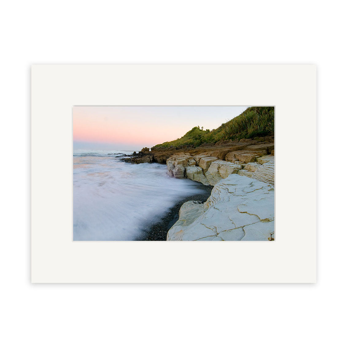 A coastal scene with rocky cliffs, waves on dark sand, and a pastel sky at sunrise or sunset, framed with a wide white mat. Punakaiki - RP201 is an exquisite New Zealand photographic print by Reuben Price.
