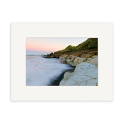 A coastal scene with rocky cliffs, waves on dark sand, and a pastel sky at sunrise or sunset, framed with a wide white mat. Punakaiki - RP201 is an exquisite New Zealand photographic print by Reuben Price.