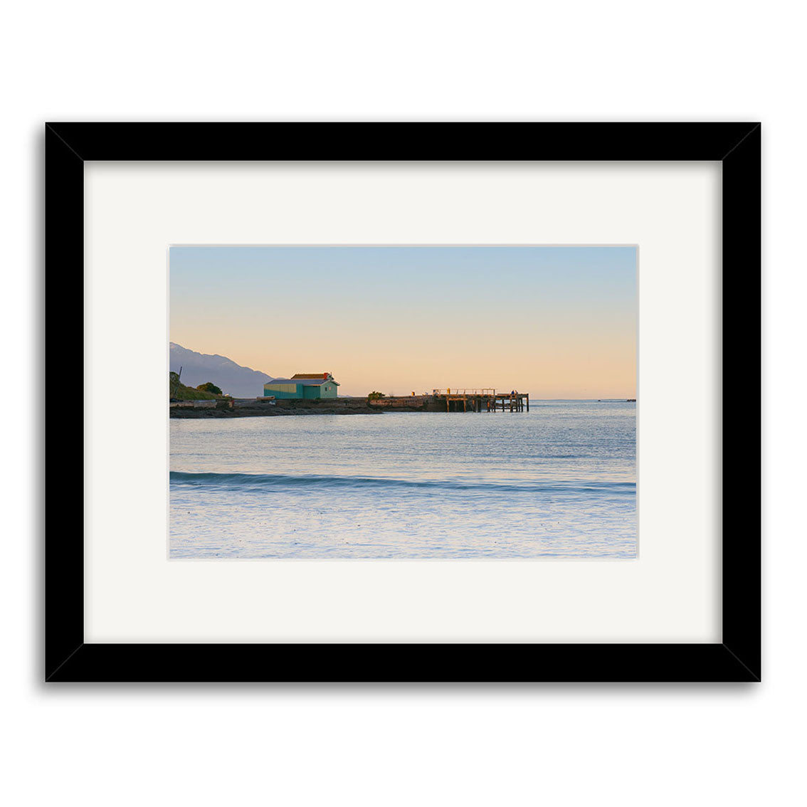 Kaikoura 1 - RP204: A framed photographic print by Reuben Price featuring a tranquil New Zealand ocean at sunset, a green building and pier extending into the water, with distant mountains under a clear sky.