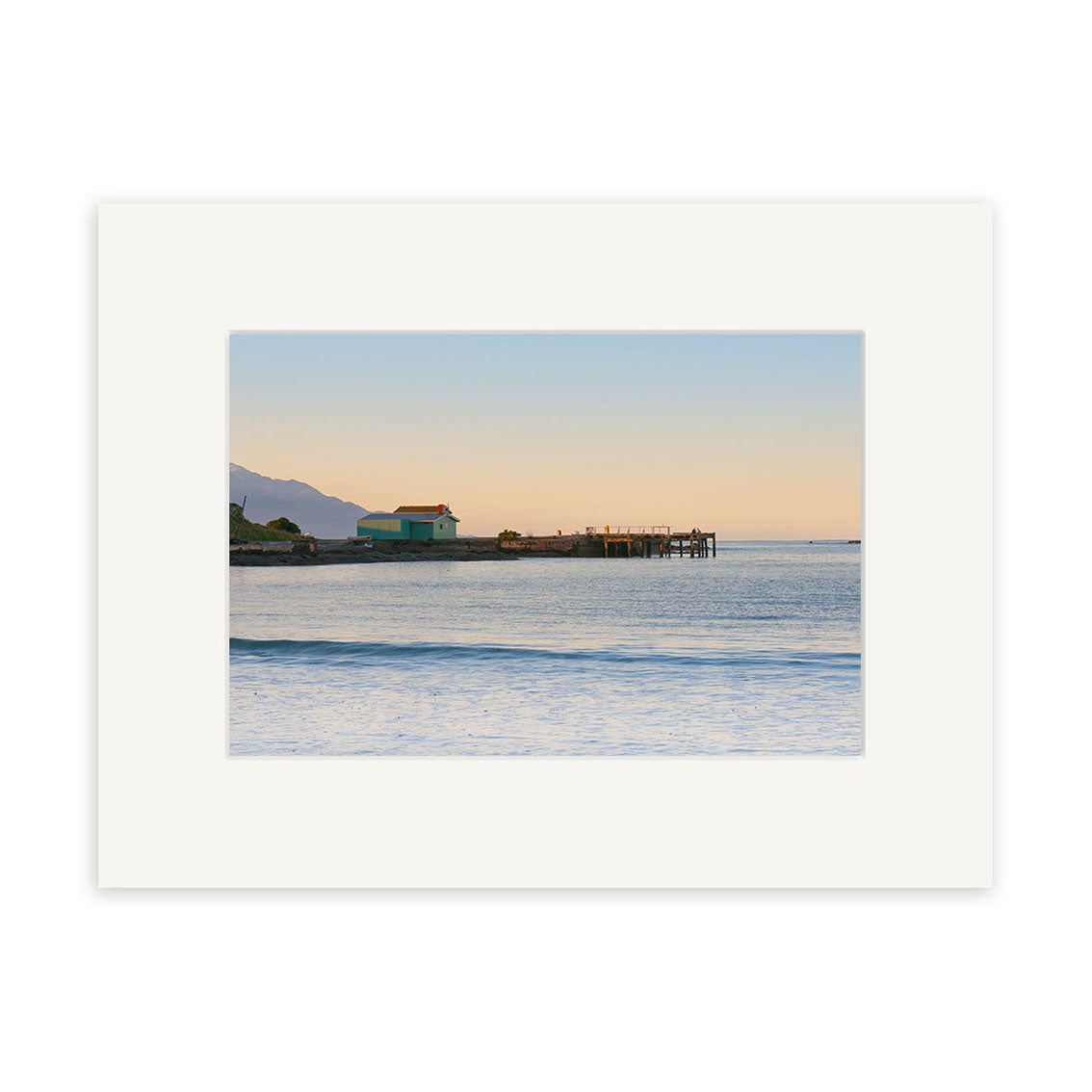 Capture the beauty of New Zealand with Kaikoura 1 - RP204: a sunset coastal scene featuring calm waves, a green building by the shore, a wooden pier over tranquil waters, and distant mountains under a clear sky—ideal for photographic prints.