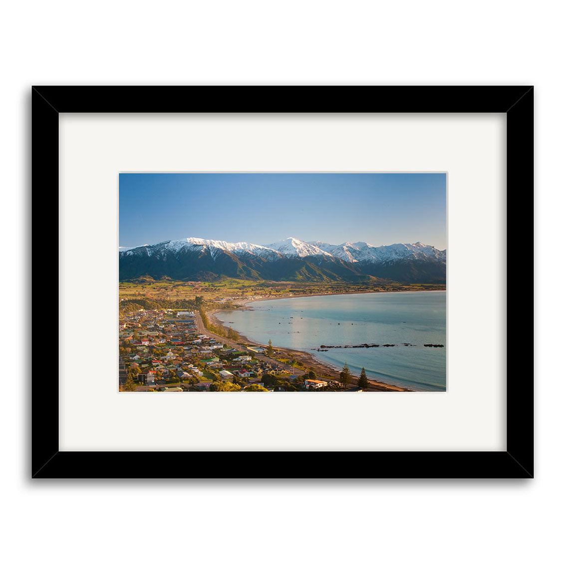 Kaikoura 2 - RP205 is a framed archival-quality photographic print by Reuben Price, featuring a coastal New Zealand town with shoreline houses, a curved bay, and snow-capped mountains beneath a clear blue sky.
