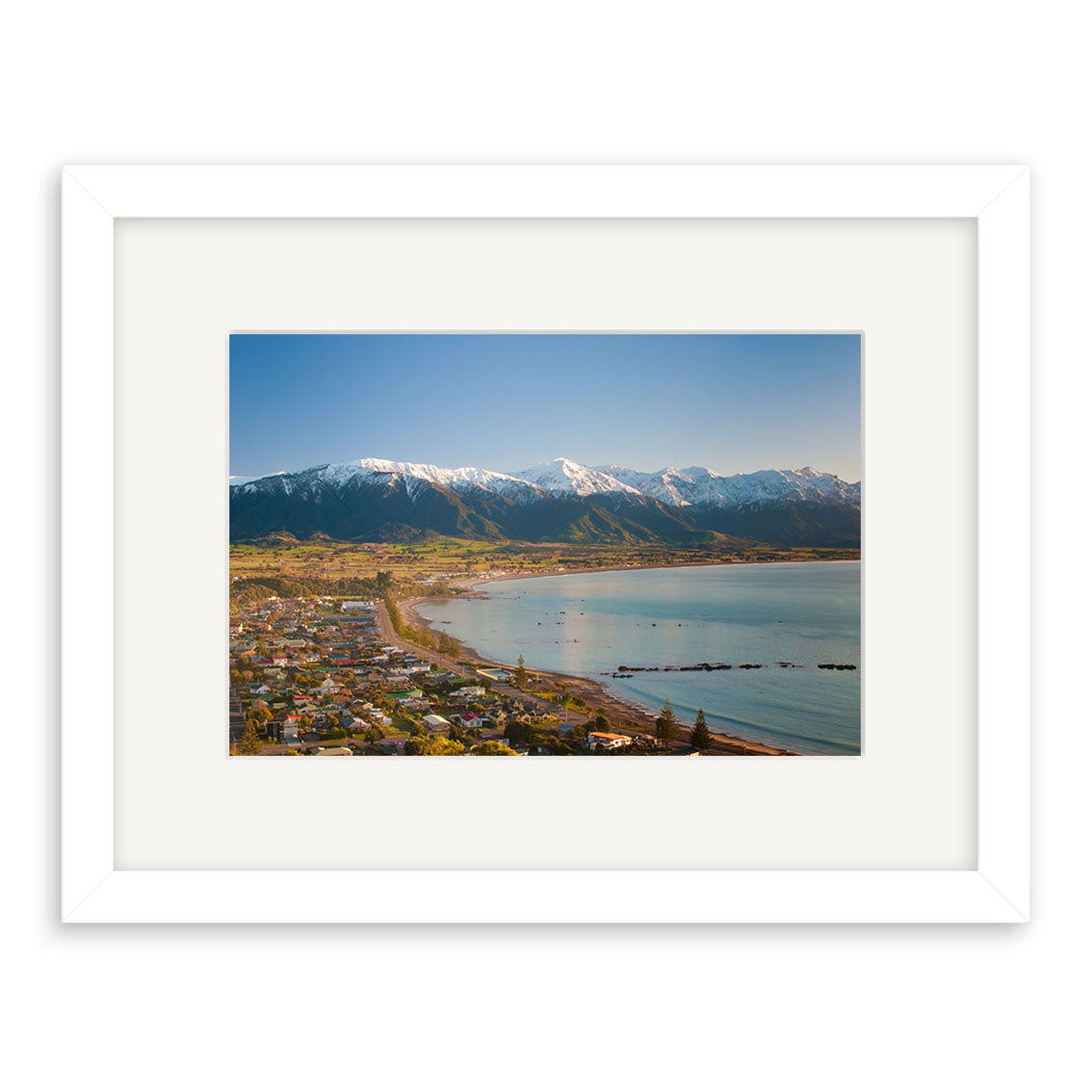 Kaikoura 2 - RP205 by Reuben Price is a framed archival-quality New Zealand print, featuring houses along a curved bay with snow-capped mountains beneath a clear blue sky.