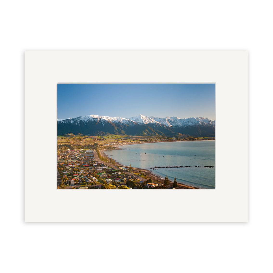 Aerial view of Kaikoura with houses, curved shoreline, and beach. Snow-capped mountains rise in the background under a clear blue sky. Kaikoura 2 - RP205 by Reuben Price is framed with a white mat for archival display.