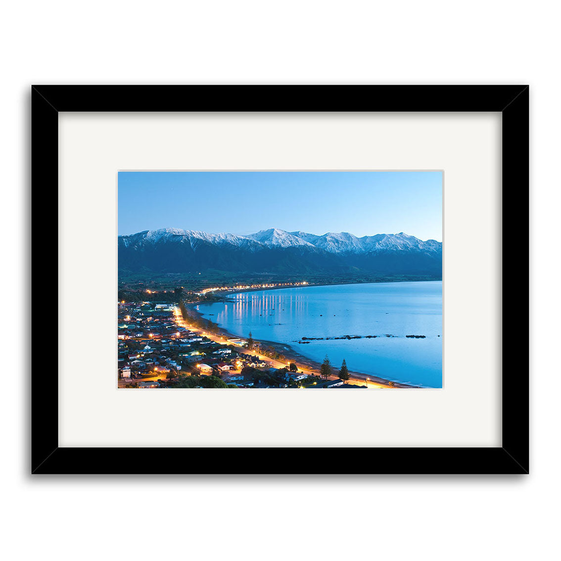 Kaikoura 3 - RP206 is a framed archival-quality New Zealand photographic print by Reuben Price, capturing city lights on the Kaikoura shoreline at dusk with calm water and snow-capped mountains under a clear sky.