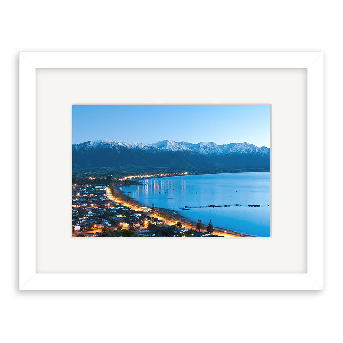 Kaikoura 3 - RP206: Archival quality New Zealand print by Reuben Price, featuring a coastal town at dusk with city lights, calm water, and snow-capped mountains under a clear blue sky.