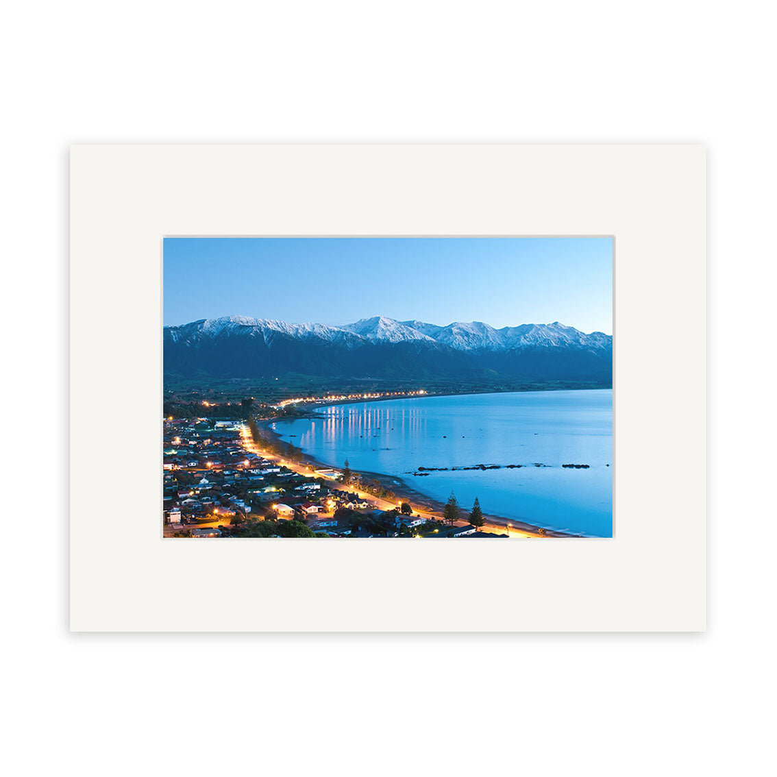 Kaikoura 3 - RP206 by Reuben Price showcases a coastal town at dusk with lit waterfront buildings, tranquil blue water, and snow-capped peaks. This New Zealand photographic print is archival quality with a wide white mat.