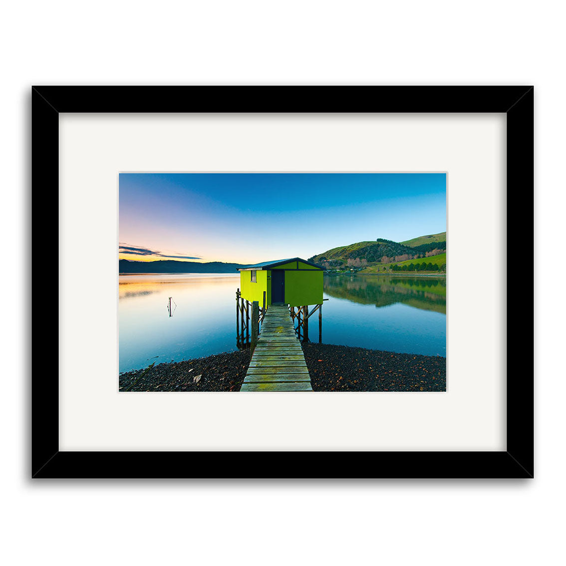 Otago Peninsula - RP207: A framed photographic print of a small green hut at the end of a wooden pier in New Zealand, over calm water at sunset. Printed on archival paper with a black frame and white mat.