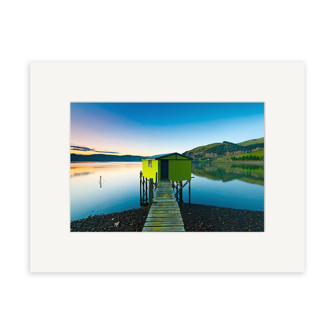 Otago Peninsula - RP207 captures a wooden walkway leading to a lime green boathouse over calm water, with hills and blue sky beyond. This New Zealand scene is presented as a photographic print on archival paper in a white matte frame.