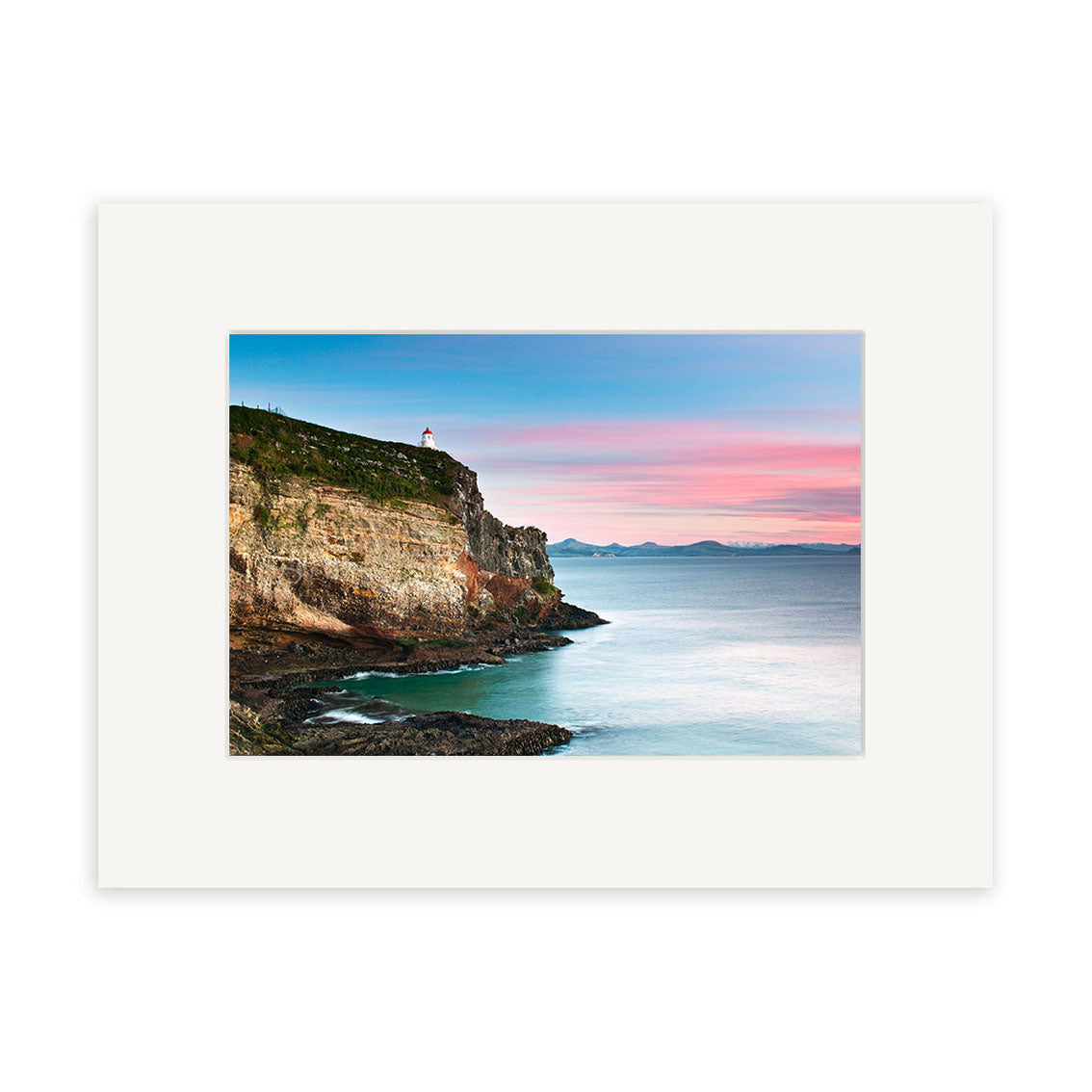 The Taiaroa Head Landscape - RP208 shows a coastal cliff with a lighthouse above calm blue water beneath a pink and blue sunset, distant mountains on the horizon. This photographic print comes matted in white and is printed on archival quality matte paper.