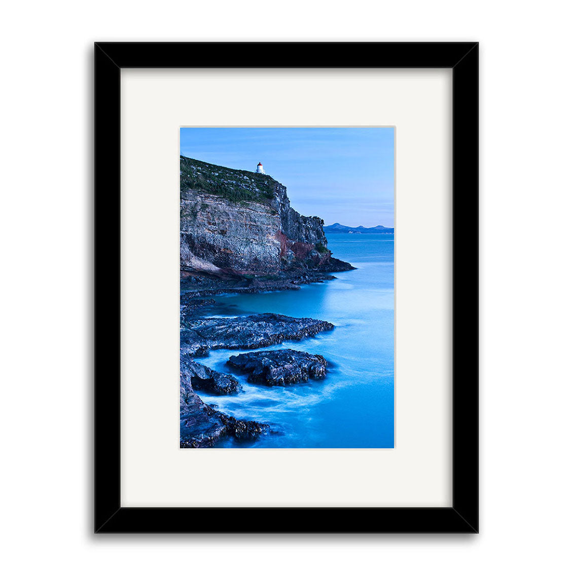 Taiaroa Head Portrait - RP209 is a framed photographic print of a coastal cliff and lighthouse in New Zealand, overlooking blue water. Presented in a black frame with white mat, its printed on archival quality matte paper for lasting beauty.