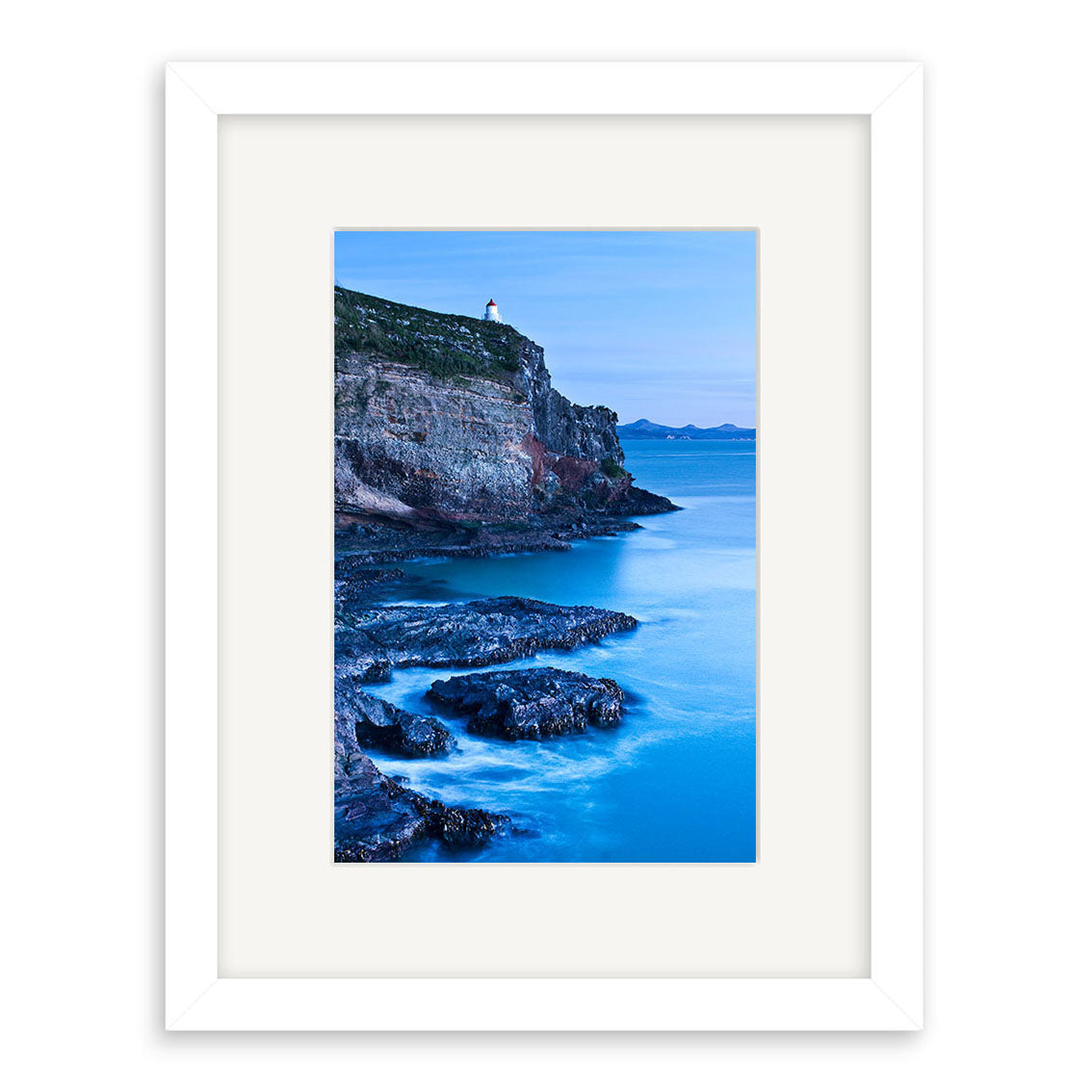 Taiaroa Head Portrait - RP209 features a rocky coastline with a lighthouse atop cliffs over calm blue water under a clear sky. Presented in a white frame with mat, its printed on archival matte paper for lasting beauty.