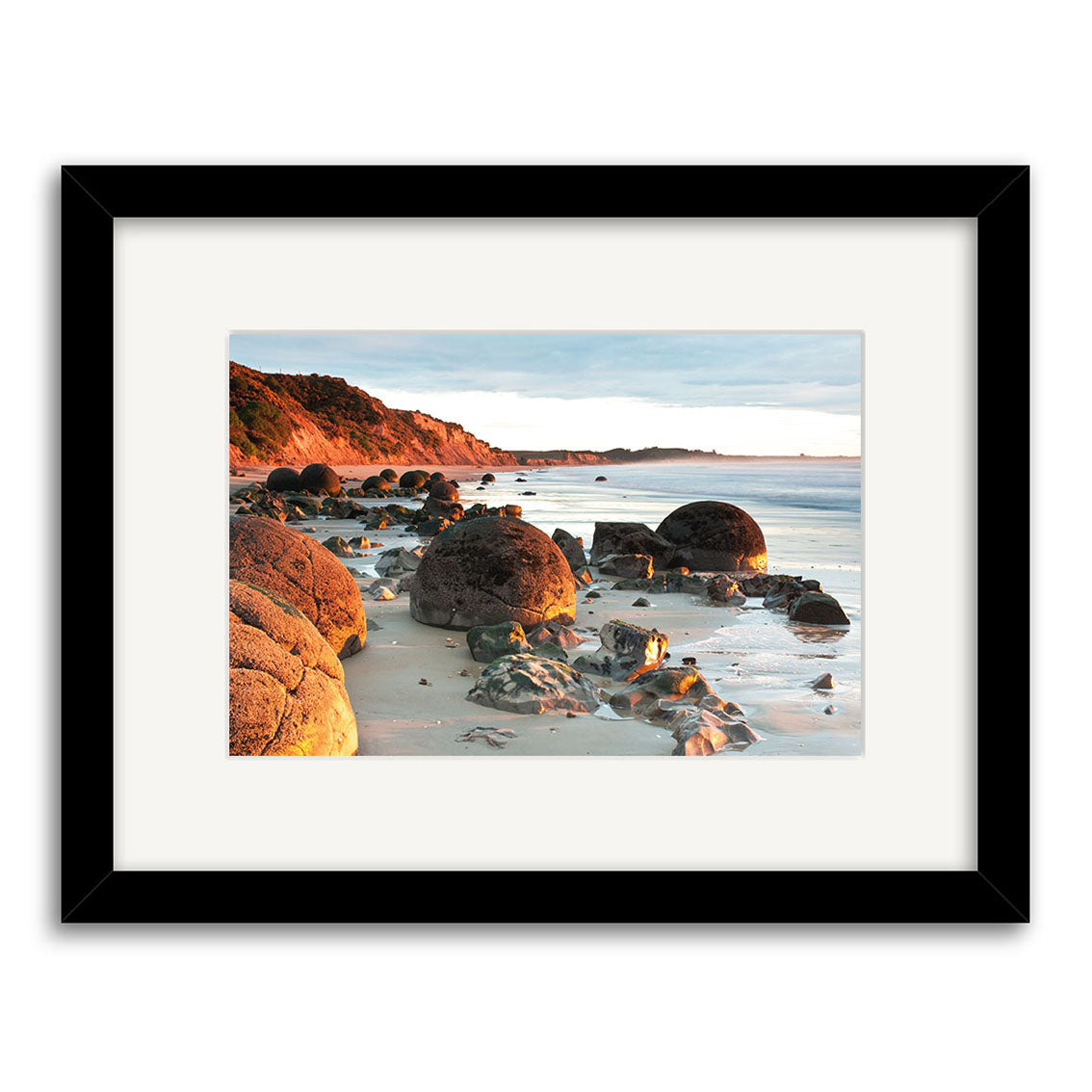 Moeraki Boulders - RP210: Archival quality framed photograph by Reuben Price, featuring large round boulders on a sandy beach with cliffs and foliage, presented with a white mat and black frame.